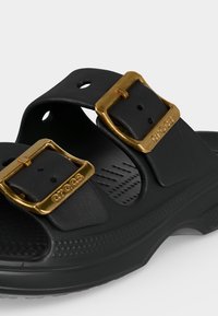 Sandale Crocs noire avec deux sangles noires réglables ornées de boucles dorées embossées du logo "Crocs".