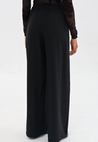 Osoba w czarnych spodniach o szerokich nogawkach z elastycznym pasem oraz przezroczystej bluzce z długim rękawem w kwiatowe wzory, stojąca na białym tle.