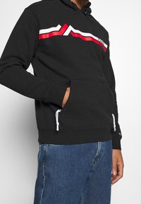 Uomo che indossa un hoodie nero con strisce geometriche rosse e bianche e jeans blu, con la mano sinistra nella tasca dell'hoodie, su uno sfondo semplice.