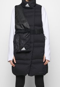 Sort polstret vest med høj krave, der har en skinnende sort stoflap, sidelomme, justerbart bælte og Adidas-logo accenter.
