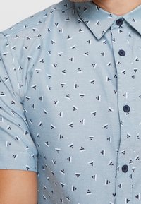 Korte mouwen shirt van lichtblauw stof met een geometrisch patroon van kleine driehoeken in donkerblauw, wit en bordeauxrood. Ontwerp met knopen aan de voorkant.