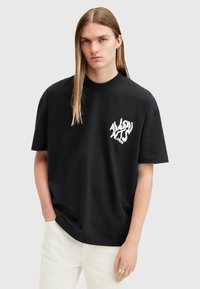 Camiseta de algodón negra con un corte relajado, que presenta una impresión gráfica blanca en el lado izquierdo del pecho. Combinada con pantalones de color claro.
