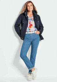 Marineblauwe gewatteerde jas met capuchon, kleurrijk geometrisch patroon top, lichtblauwe jeans en witte sneakers. Voorzien van een riem en opgerolde mouwen.