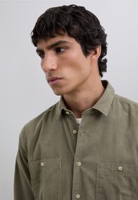 Chemise en coton vert olive avec un col boutonné, deux poches poitrine et un tissu texturé. Présente des boutons beiges et des détails de couture subtils.