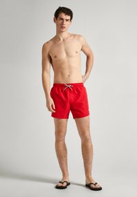 Sarkanie peldbikses ar elastīgu jostu un auklu. Materiāls izskatās viegls un gluds. Apvienots ar melniem flip-flop apaviem.