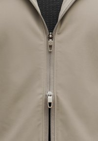 Helle beigefarbene Zip-Jacke mit glatter Textur, die einen Doppel-Reißverschluss und detaillierte Nähte an der Vorderseite aufweist, vor einem dunkel gemusterten Innenfutter.