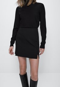 Haut à manches longues noir associé à une mini-jupe noire avec une fente sur le côté. Le mannequin porte également des bottes noires jusqu'aux genoux, mettant en valeur un design élégant.