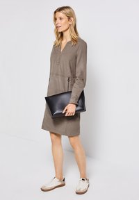 Taupe Langarmkleid mit V-Ausschnitt, zwei seitlichen Taschen und tailliertem Bund. Kombiniert mit einer schwarzen Clutch und weißen Sneakers.