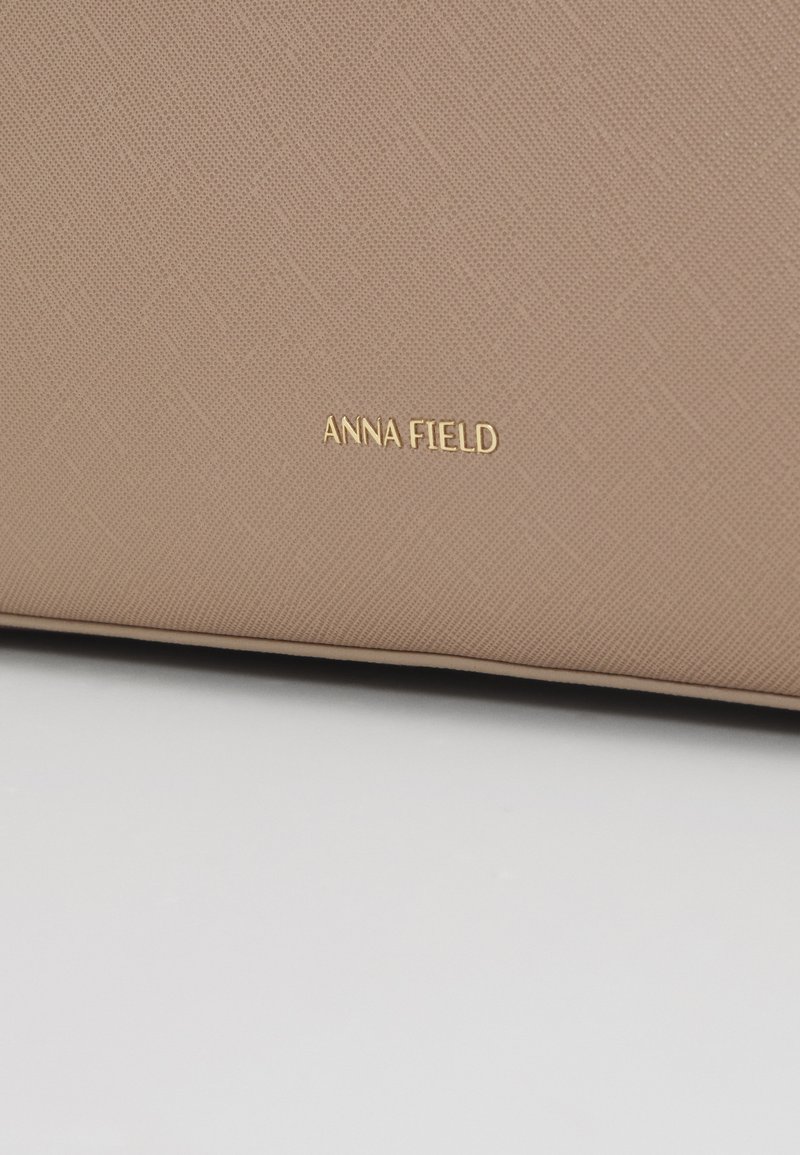 Bolso beige texturizado con un sutil patrón geométrico, que presenta un logo dorado "ANNA FIELD". Material suave con una forma estructurada.