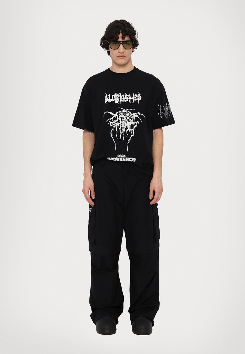 Sort oversized t-shirt med hvide grafiske print, parret med sorte cargo-bukser med store lommer. Modellen bærer solbriller med grønne nuancer.