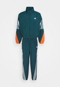 Ensemble de survêtement Adidas vert foncé avec veste zippée intégrale et pantalon, présentant des bandes blanches et des accents orange sur les manches.