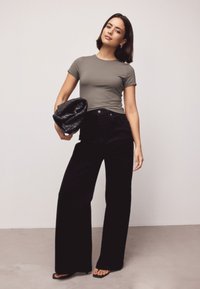 Kvinde stående med sort vævet clutch, iført tætsiddende grå kortærmet top, højttaljede, vidbenede sorte bukser og sorte åbne hæle.