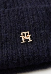 Căciulă tricotată de culoare bleumarin, cu un pin metalic auriu ce prezintă litere interconectate. Țesătura are o suprafață texturată, cu o formă profilată.