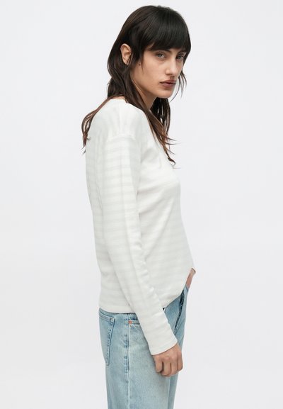 Chemise blanche à manches longues rayée avec une texture douce et une coupe décontractée, associée à un jean bleu clair, prise de profil sur un fond uni.
