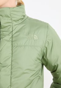 Svetlozelená izolačná bunda s vysokým golierom, s vyšívaným logom The North Face na hornej ľavej strane. Hladká, matná textura.
