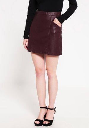 Femme portant une mini-jupe en cuir bordeaux avec une poche, un haut noir à manches longues, et des sandales noires à talons hauts avec bride à la cheville.