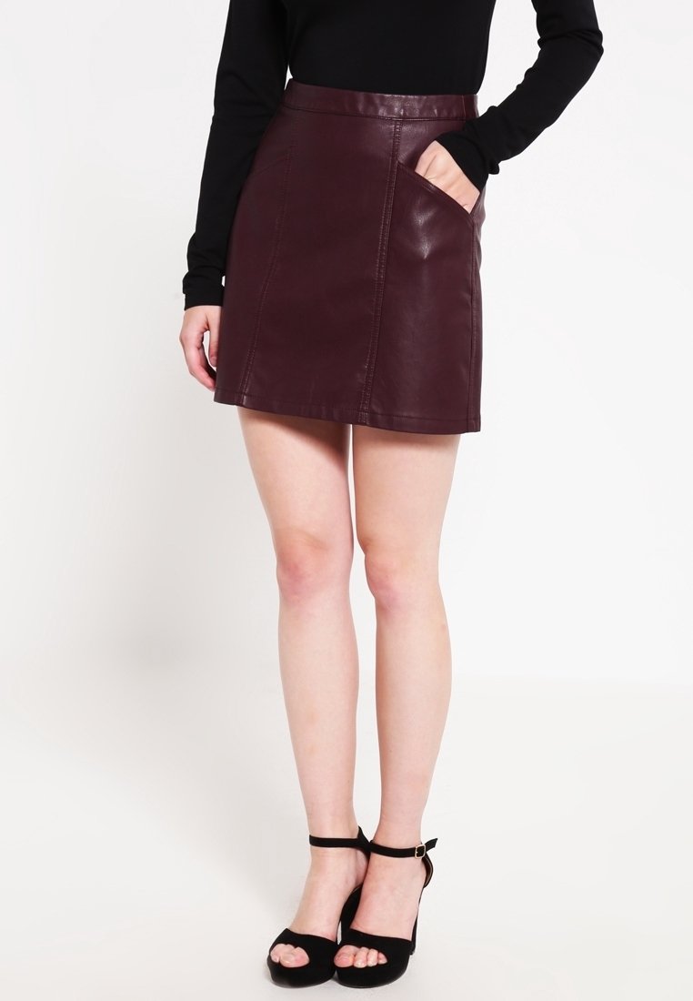 Femme portant une mini-jupe en cuir bordeaux avec une poche, un haut noir à manches longues, et des sandales noires à talons hauts avec bride à la cheville.