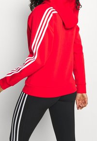 Sudadera roja ajustada, con franjas horizontales blancas en las mangas y un bolsillo tipo canguro. Combinada con leggings negros que tienen franjas blancas.