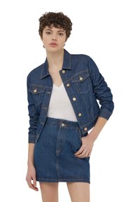 Giubbotto di jeans blu scuro, con bottoni color oro, due tasche sul petto e un design cropped, indossato sopra un top bianco a V e una gonna di jeans.