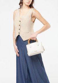 Gilet beige senza maniche con bottoni, abbinato a pantaloni ampi blu navy. Il modello tiene una piccola borsa bianca con manico in bambù e dettagli dorati.