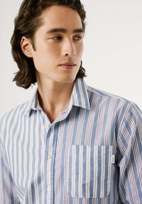 Camisa de algodón ligera con rayas verticales en azul, blanco y rojo, con cuello de botón y un bolsillo en el lado izquierdo del pecho.