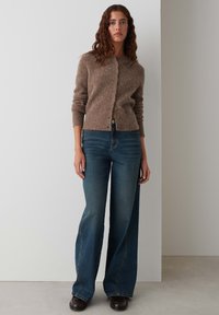 Bruine gebreide cardigan met knopen, gecombineerd met hoog-waisted blauwe flare jeans met zichtbare stiksels en een gladde textuur.