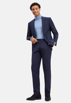 Junger Mann lächelnd, trägt einen marineblauen Anzug mit einem hellblauen Rollkragenpullover und schwarzen Schuhen, steht mit einer Hand in der Tasche.