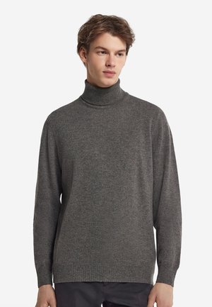 Ung mand iført en almindelig grå rullekravesweater og mørke bukser, ser let til siden mod en hvid baggrund.