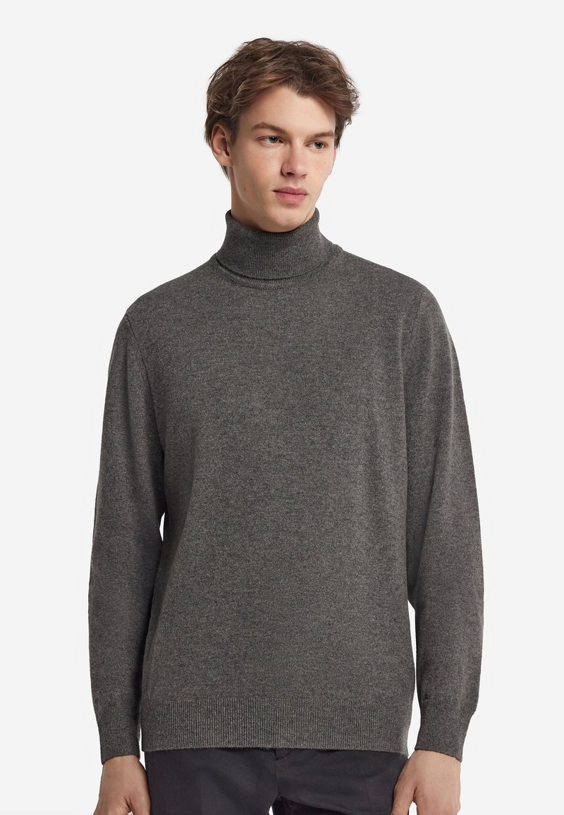 Junger Mann mit einfachem grauem Rollkragenpullover und dunkler Hose, schaut leicht seitlich vor weißem Hintergrund.