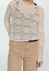 Jasnobeżowy sweter dzianinowy z teksturowanym wzorem ażurowym; ma długie rękawy i okrągły dekolt. Widoczny brązowy top pod spodem.