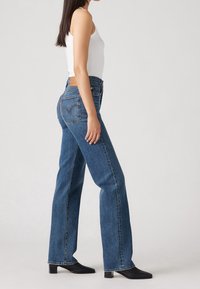 Kvinna som bär högmidjade raka blåjeans, vit ärmlös topp och svarta klackstövlar, stående sida vid sida mot en enhetlig bakgrund.