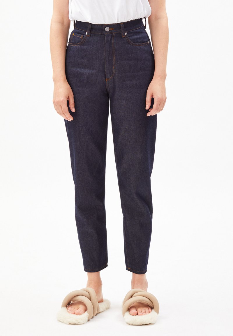 Jean taille haute en denim foncé avec des surpiqûres orange, coupe droite et poches latérales, associés à des pantoufles moelleuses de couleur claire.