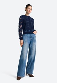 Cardigan in maglia blu navy con ricamo floreale e maniche trasparenti, abbinato a jeans a gamba larga azzurri e tacchi marroni a punta.