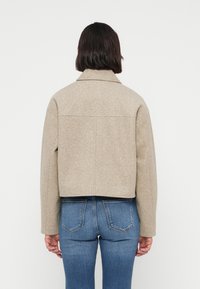 Μπεζ κοντό μπουφάν με γιακά, από υφασμένο ύφασμα. Διαθέτει μακριά μανίκια και καθαρή εφαρμοστή κοπή, συνδυασμένο με μπλε τζιν παντελόνι.