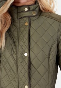 Gequiltte, olijfgroene jas met gouden drukknoopjes en een bruine bies, voorzien van een hoge kraag en verticale ribgebreide schouderaccenten.