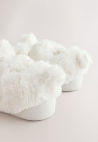 Chaussons blancs et moelleux avec des oreilles d’ours sur le dessus, sur un fond neutre, inclinés pour montrer la vue arrière et latérale.