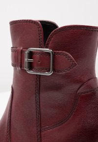 Bottine en cuir rouge foncé avec coutures visibles et bride à boucle rectangulaire argentée près du bord supérieur.