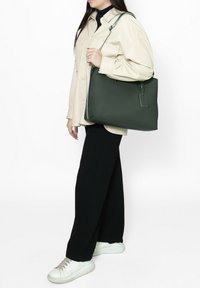 Sac fourre-tout en cuir vert avec une texture lisse, des poignées supérieures et une forme rectangulaire, accompagné d'une étiquette amovible.