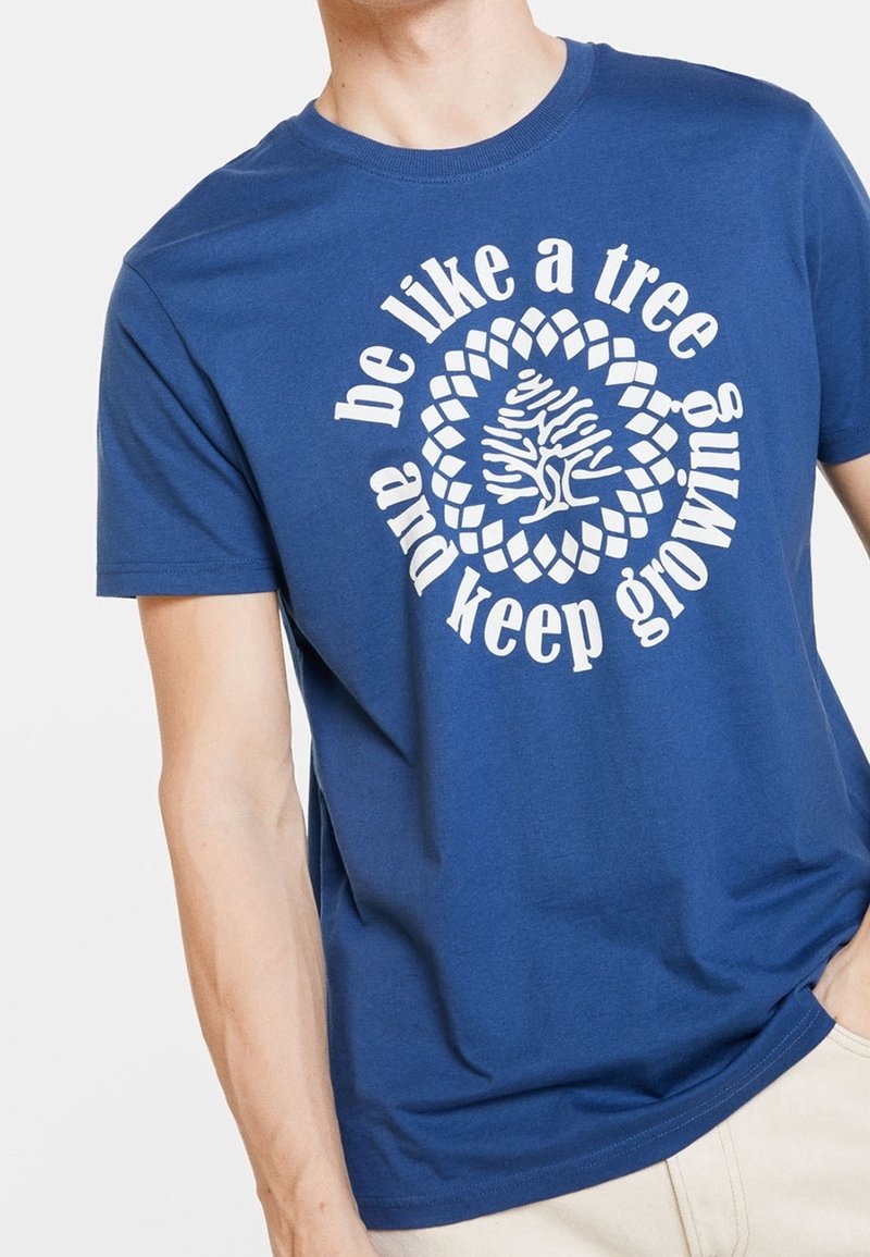 T-shirt bleu avec un texte circulaire blanc « sois comme un arbre et continue de grandir » entourant un graphisme stylisé d'un arbre blanc sur le torse d'une personne.