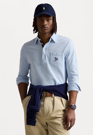 Light blue long-sleeve polo shirt with a chest pocket, navy sweater tied at the waist, khaki pants, and a navy baseball cap.