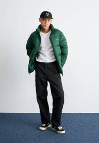 Giacca a vento verde, t-shirt bianca, pantaloni neri e cappellino nero. Le caratteristiche includono texture trapuntata, vestibilità ampia e accenti a contrasto.