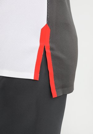 Harmaa- ja valkoinen urheilupaita, jossa punaiset yksityiskohdat; siinä on verkkokangasrakenne, sivuhalkiot ja moderni leikkaus parannetun ilmanvaihdon takaamiseksi.