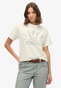 Cremefarvet t-shirt med korte ærmer og teksten "SUPER DRY ATHLETIC" trykt i grå, med en afslappet pasform og blød tekstur, sammen med stribede bukser og et brunt bælte.