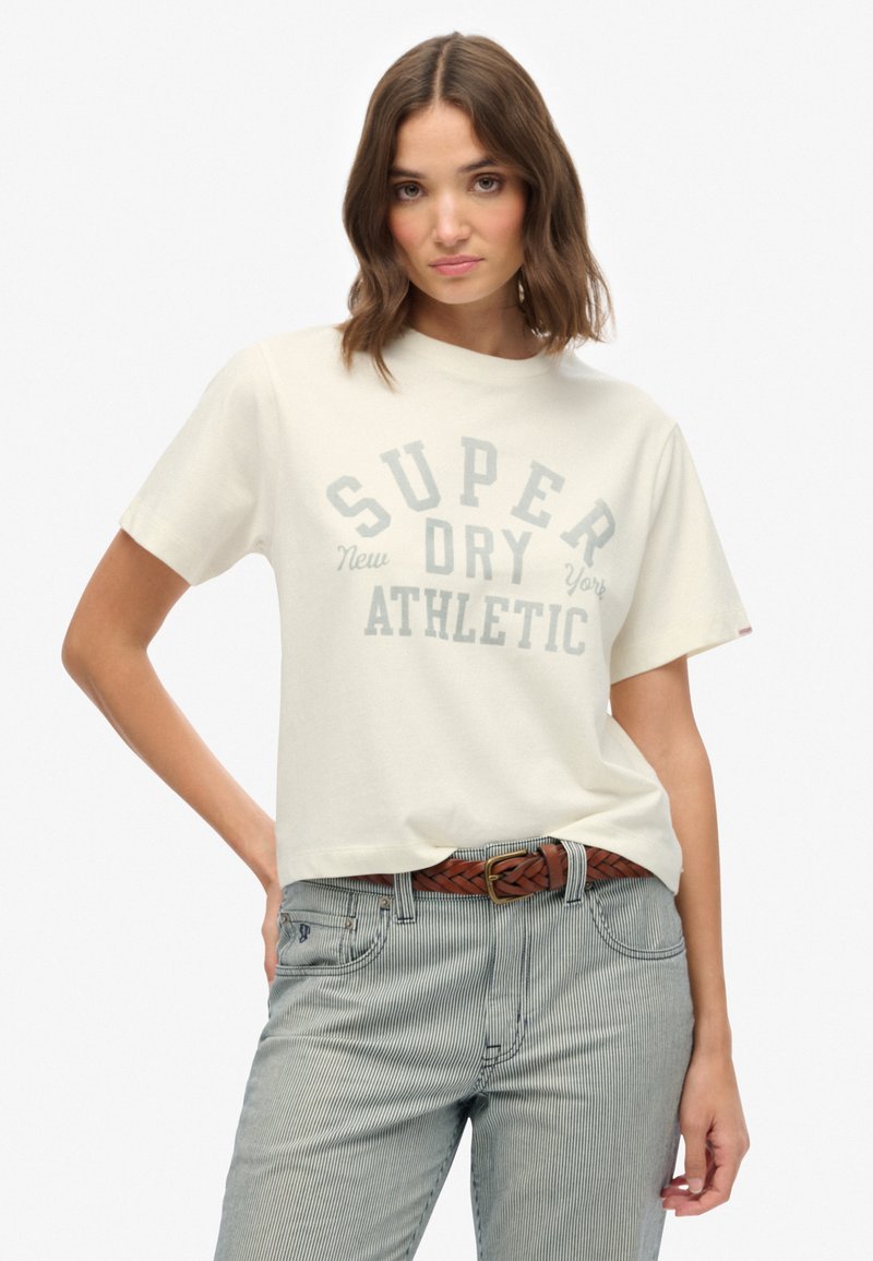Cremefarvet t-shirt med korte ærmer og teksten "SUPER DRY ATHLETIC" trykt i grå, med en afslappet pasform og blød tekstur, sammen med stribede bukser og et brunt bælte.