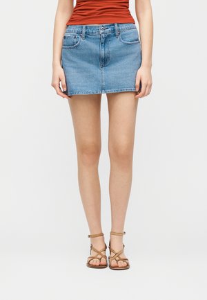 Femme portant une mini-jupe en denim bleu, un haut orange et des sandales plates à brides marron, debout devant un fond blanc uni.