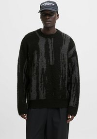Ein schwarzer Mann trägt einen schwarz-grau gemusterten Strickpullover, schwarze Hosen und eine schwarze Mütze mit weißem Text vor einem einfarbigen Hintergrund.