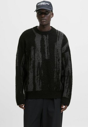 Black man wearing a black and gray patterned knit sweater, black pants, and a black cap with white text on a plain background.