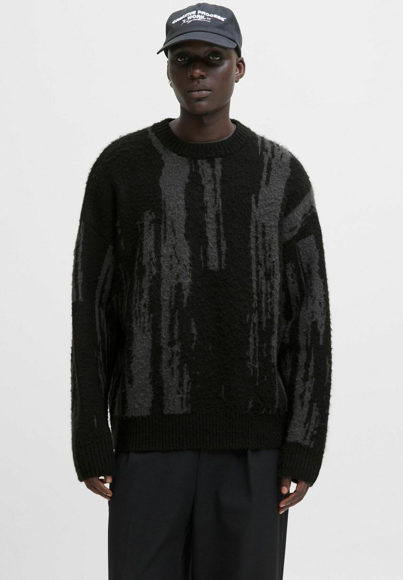 Ein schwarzer Mann trägt einen schwarz-grau gemusterten Strickpullover, schwarze Hosen und eine schwarze Mütze mit weißem Text vor einem einfarbigen Hintergrund.