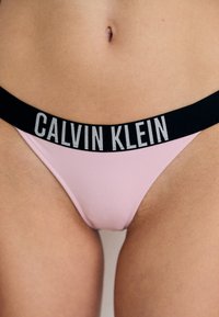 Růžové tangové spodní prádlo s černým elastickým pasem, na kterém je bílé logo "CALVIN KLEIN". Hladká textura, minimalistický design.