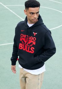 Felpa nera con cappuccio, caratterizzata da un logo rosso "Chicago Bulls" e un'immagine di toro, con cappuccio regolabile e tasca frontale. Indossata sopra una maglietta bianca.